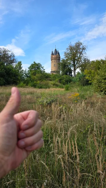 A thumbs-up in a meadow, a tower in the background.